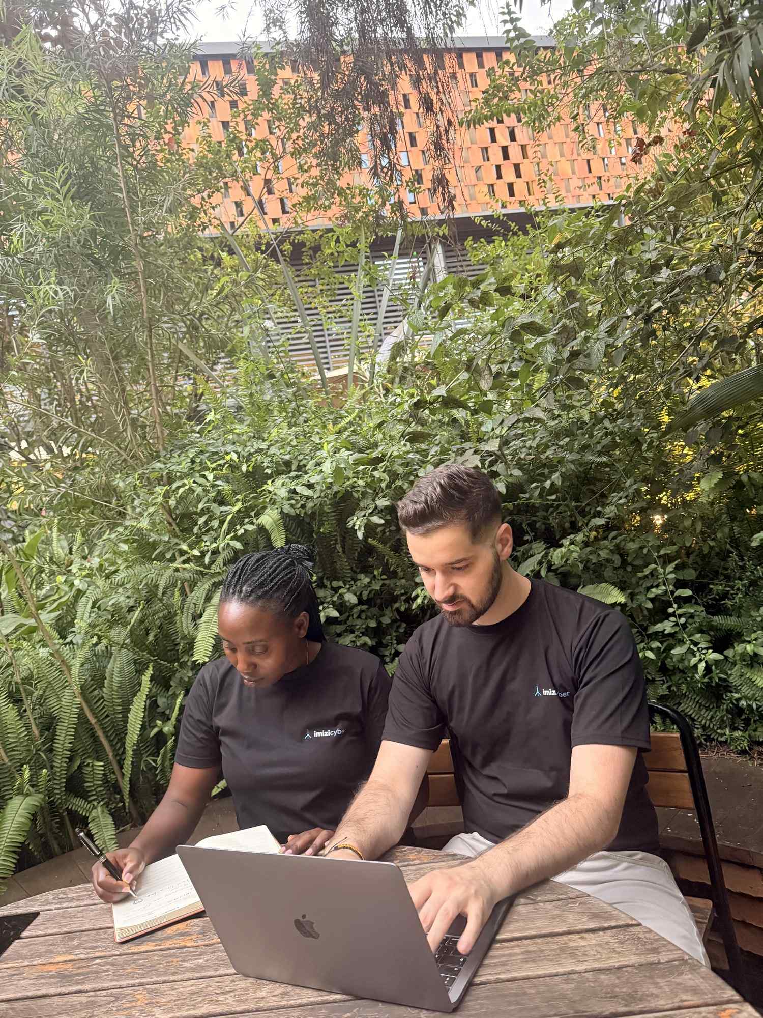 The IMIZI Cyber team working on a laptop and notebook in the garden at Norrsken House, Kigali.
